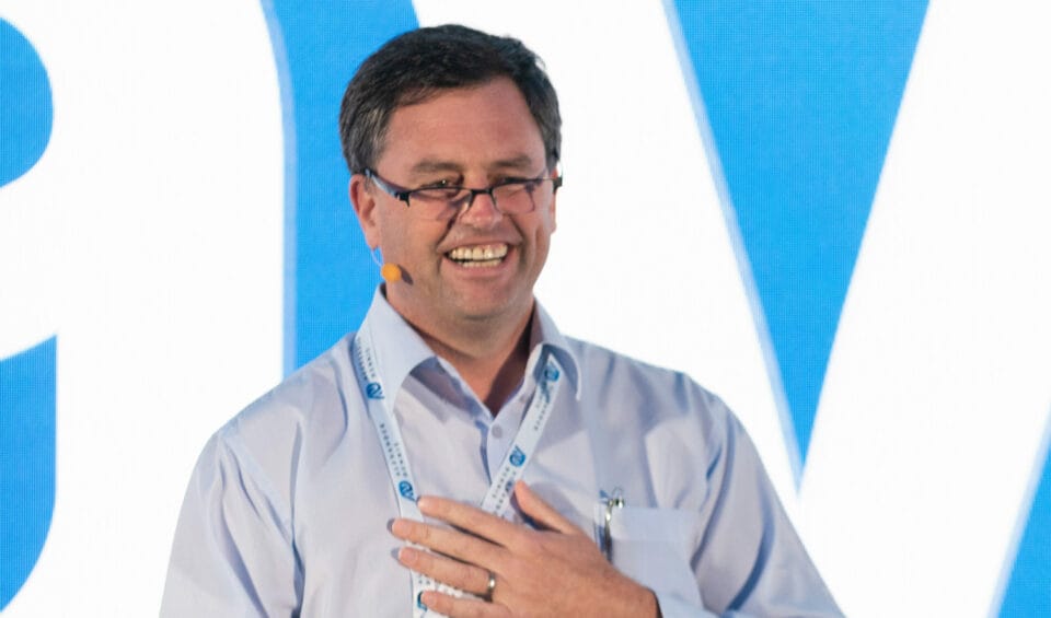 A middle-aged white man speaks in front of a large screen. He is wearing a lanyard with the Alexander Dennis logo and has one hand placed across his chest.