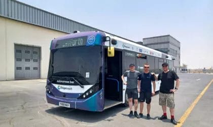 Three men wearing casual clothing for hot weather stand next to a single deck bus with heavily tinted windows in apparent strong sunshine.
