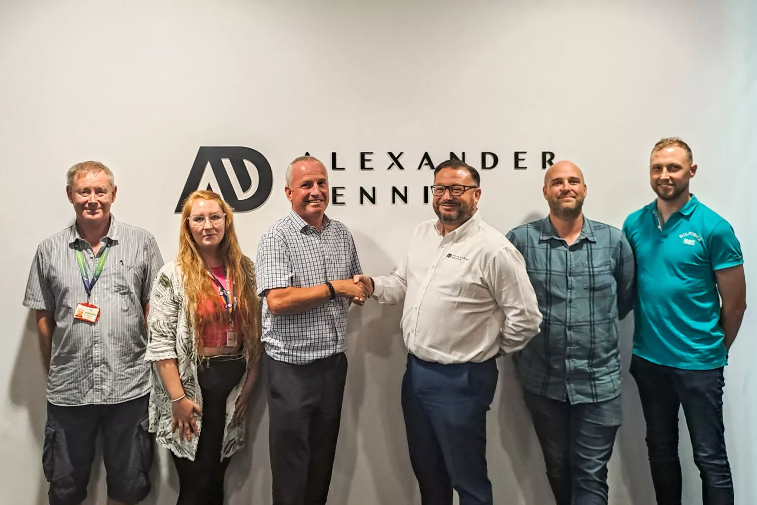 Six white people stand in front of a white wall with the Alexander Dennis logo on it. The two men in the centre shake hands.