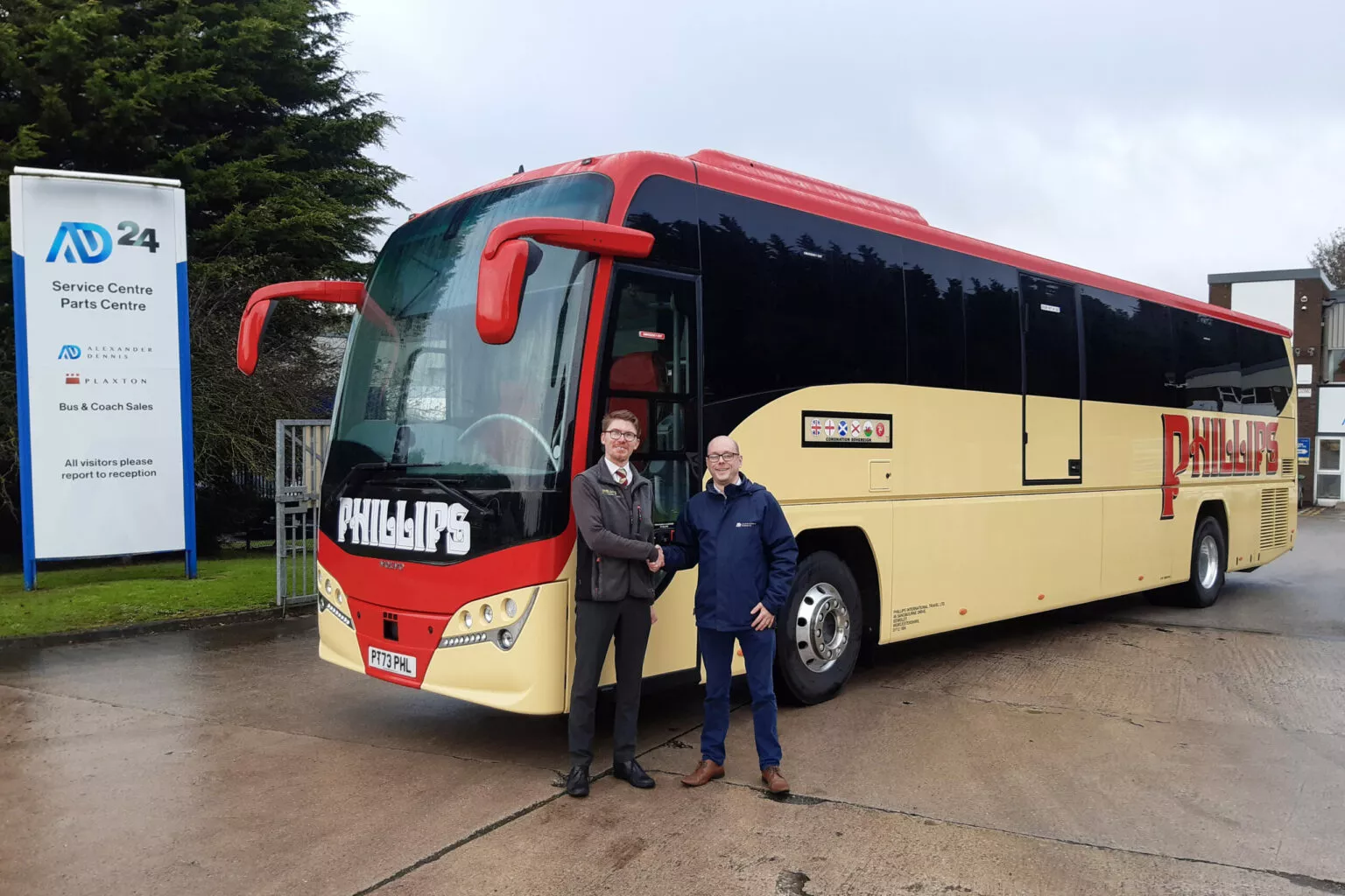 Two men stand in front of a cream and red coach, shaking hands.