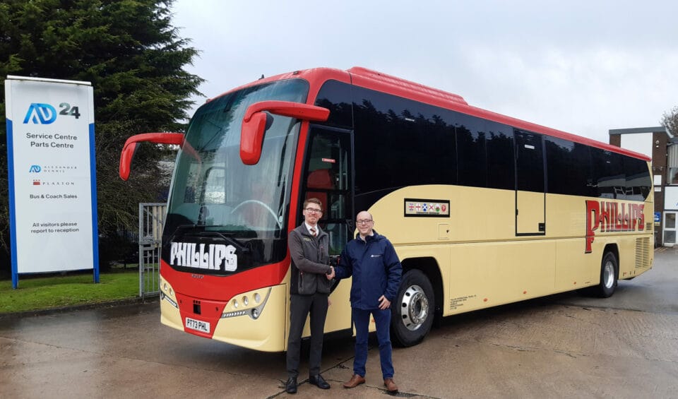 Two men stand in front of a cream and red coach, shaking hands.