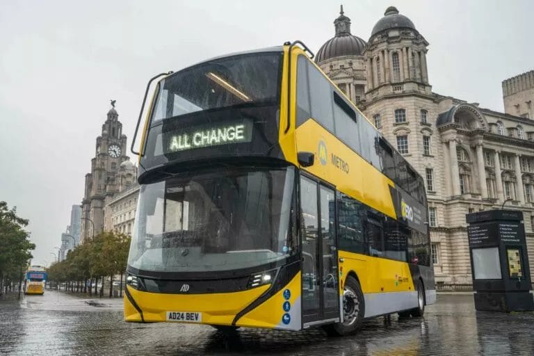 An Alexander Dennis Enviro400EV in the colours of the Liverpool City Region's Metro network stands in front of the Three Graces buildings on Liverpool's Pier Head.