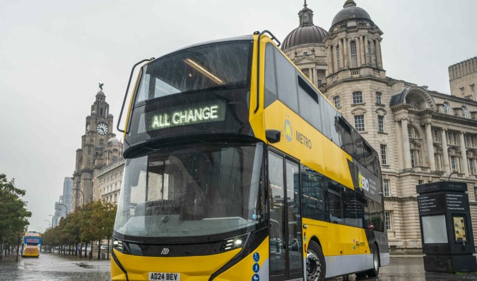 An Alexander Dennis Enviro400EV in the colours of the Liverpool City Region's Metro network stands in front of the Three Graces buildings on Liverpool's Pier Head.